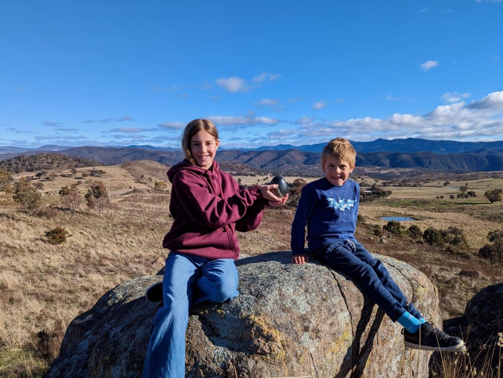mora and sven with the travelling sculputre in their hands, snowy mountains, nsw in the background